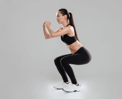 Full length of concentrated young woman athlete doing squats over gray background